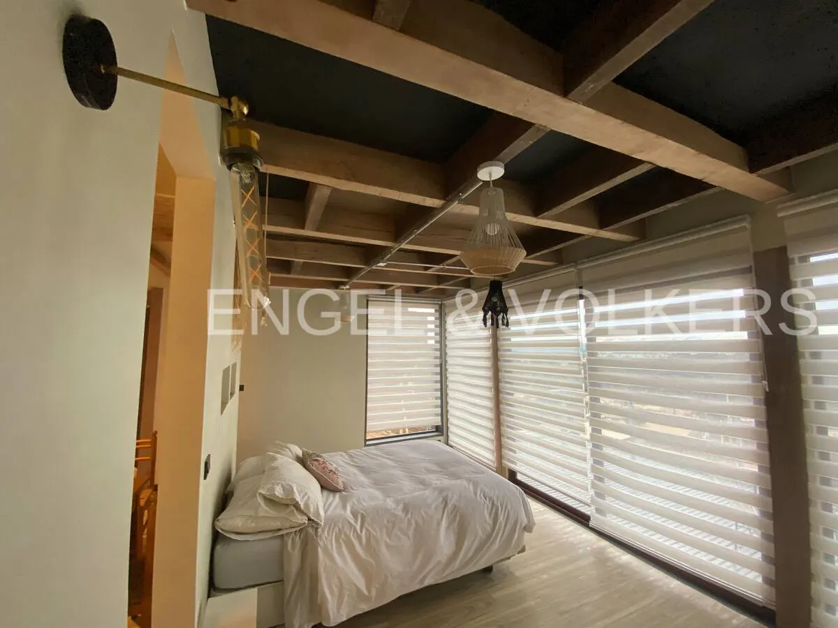 Modern bedroom with a white bed, exposed wooden ceiling beams, and large windows covered by horizontal blinds.