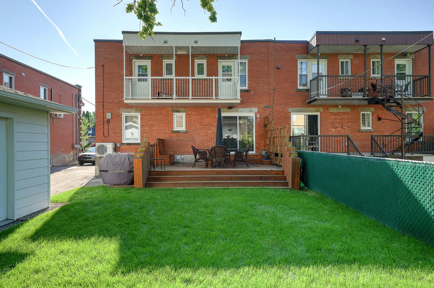Backyard view of a red brick building with a wooden deck, table, chairs, and green lawn. Balconies and a spiral staircase are visible.