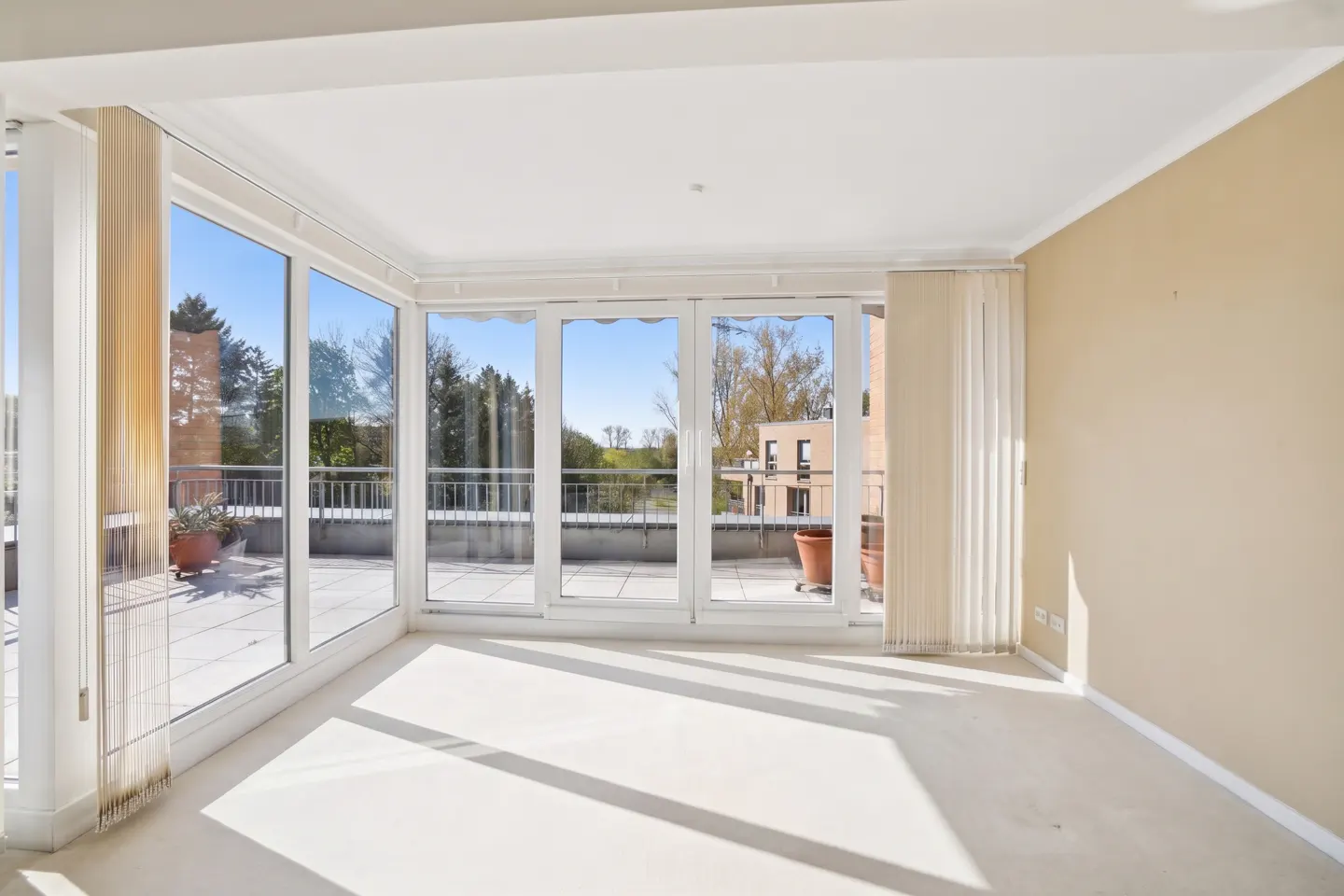 Bright, empty room with beige walls and white carpet. Large windows lead to a balcony with potted plants and a view of trees and buildings.