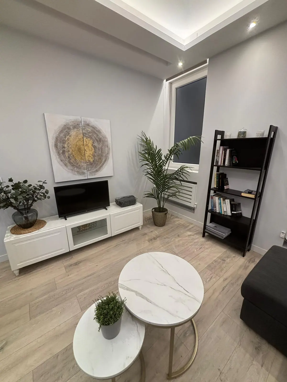 Living room with white walls, light wood floors, and modern decor. A white media console holds a TV and artwork. Two round marble tables sit in front of a dark gray sofa.