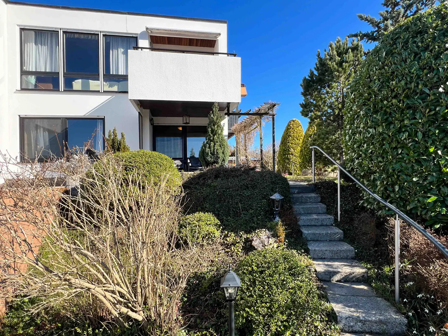 Exterior view of a modern white house with a balcony, surrounded by lush greenery and a stone staircase leading up to the entrance.