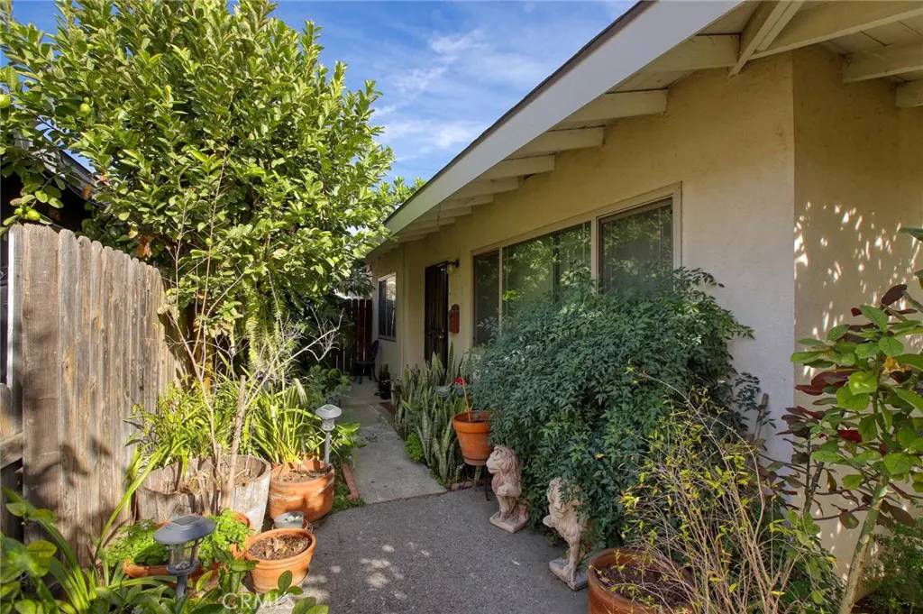A tan house with a white roof and windows is surrounded by greenery and potted plants. A narrow walkway leads to the front door.