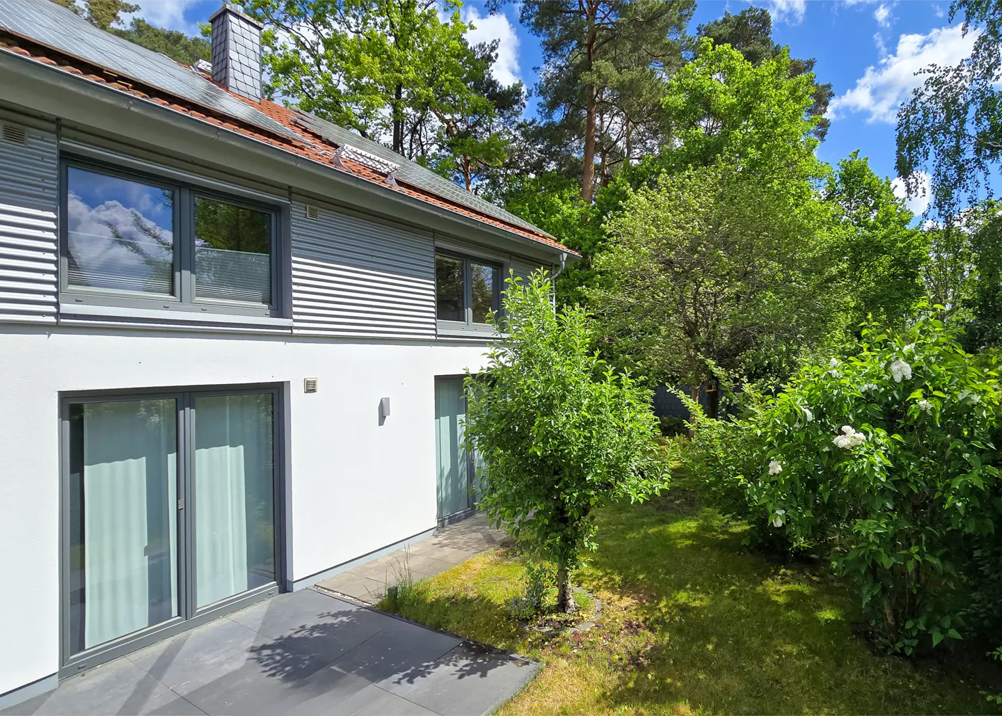 Modern two-story house with white and grey siding, large windows, and a patio overlooking a lush green garden and trees.