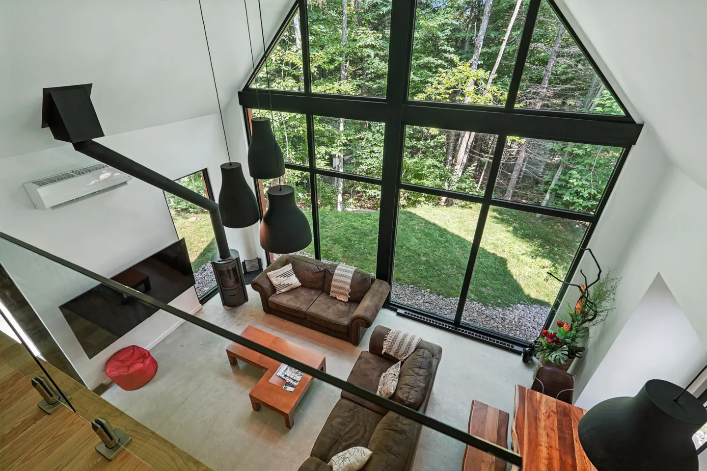 A high-angle view of a modern living room with a large A-frame window overlooking a forest. Two brown sofas, a wood stove, and black pendant lights are visible.