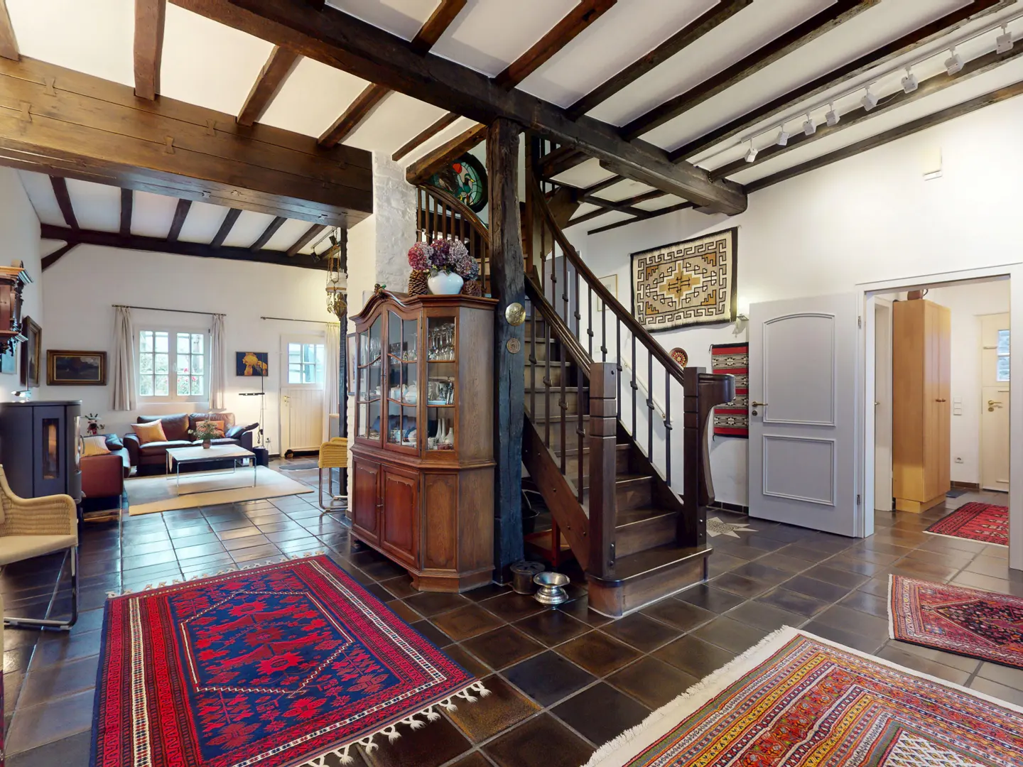 Interior view of a home's foyer with dark wood beams, staircase, and tiled floor with colorful rugs. A living room is visible in the background.