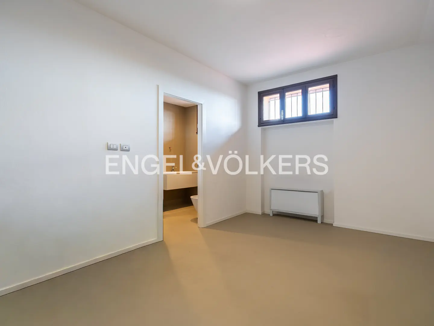 Bright, empty room with beige floor, white walls, and a brown-framed window. An open doorway reveals a bathroom with a white sink. A white radiator sits under the window.