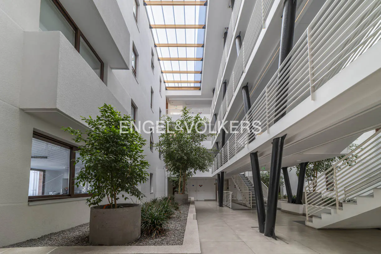 Interior view of a white building with a glass roof, plants, and walkways with white railings. Black pillars support the walkways. The Engel & Volkers logo is visible.