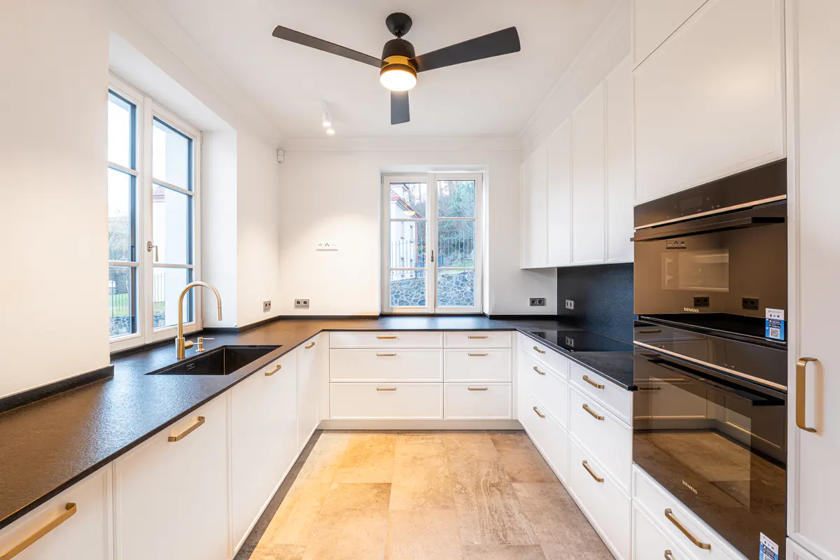 Bright kitchen with white cabinets, black countertops, and gold hardware. A black ceiling fan hangs above. Natural light streams through a window.
