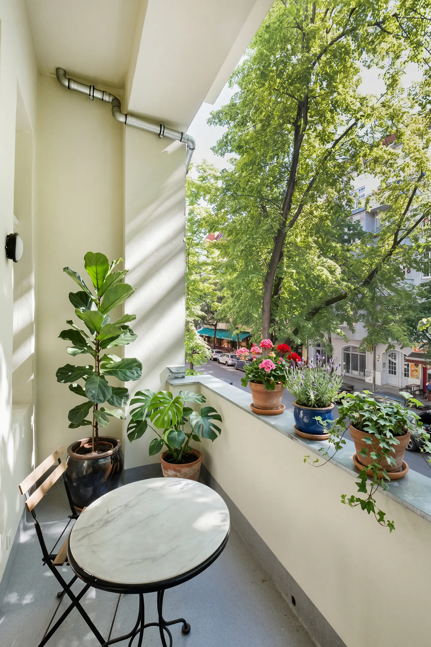 A balcony with a round marble table, two chairs, and several potted plants overlooking a tree-lined street.