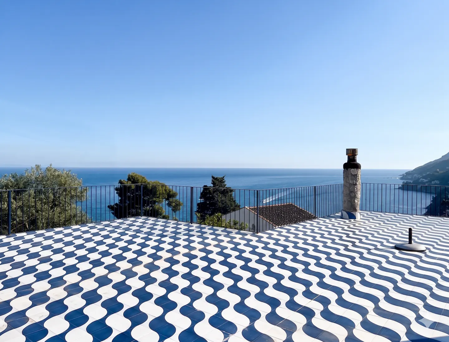 View from a rooftop terrace with blue and white wavy tiles, black railing, and ocean view under a clear blue sky.