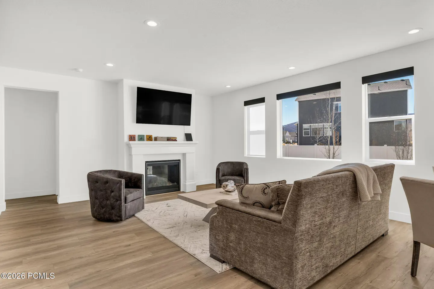 Bright living room with wood floors, white walls, and a fireplace. A brown sofa and chairs sit on a light rug. Windows show a house and mountains.