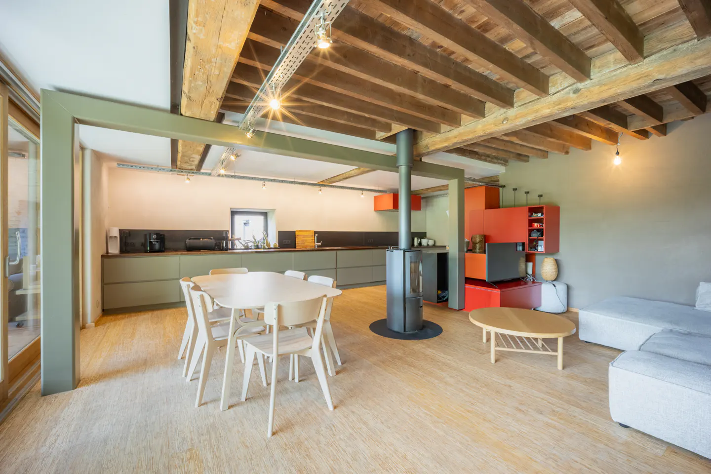 Open-plan living area with wood-beamed ceiling, dining table, modern wood stove, and orange cabinetry in a bright room.