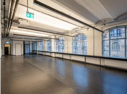 An empty commercial space with large windows, a dark floor, and overhead lighting. An exit sign is visible.