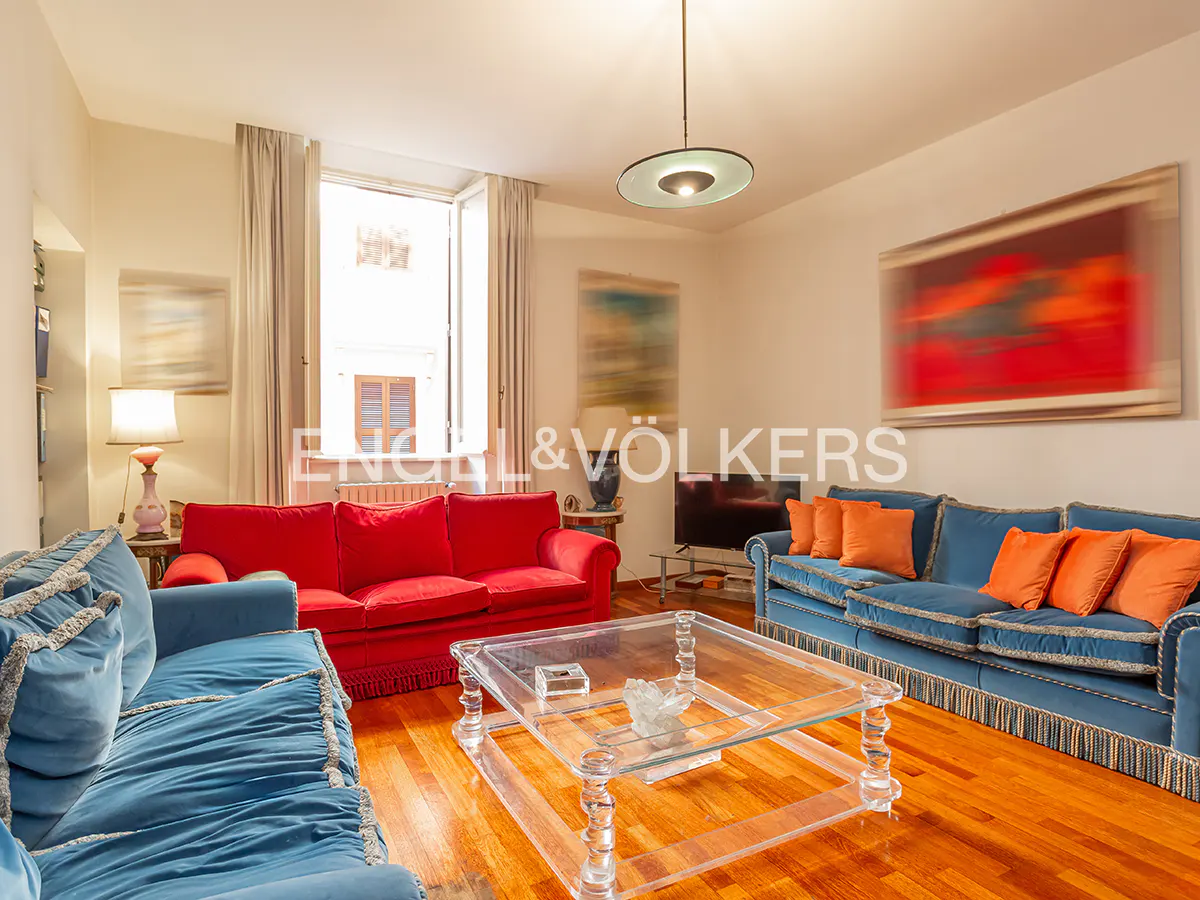Living room with red and blue sofas, a clear coffee table, and hardwood floors. A window is in the background.