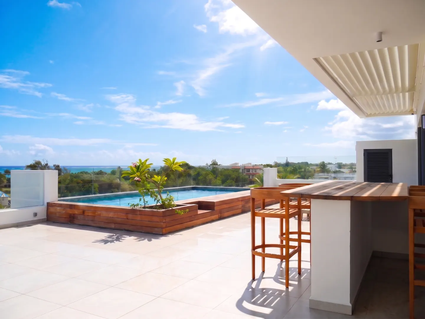 Rooftop patio with a small pool, wooden bar with stools, and ocean view under a bright blue sky.