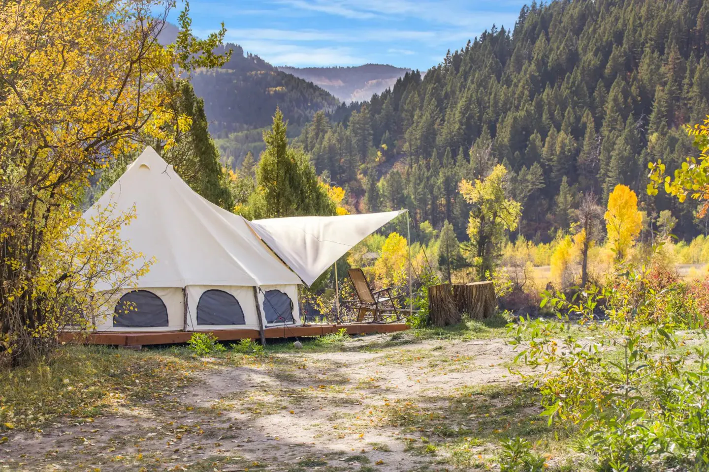 A white bell tent on a wooden deck sits in a forest clearing with autumn trees, mountains, and a blue sky in the background.