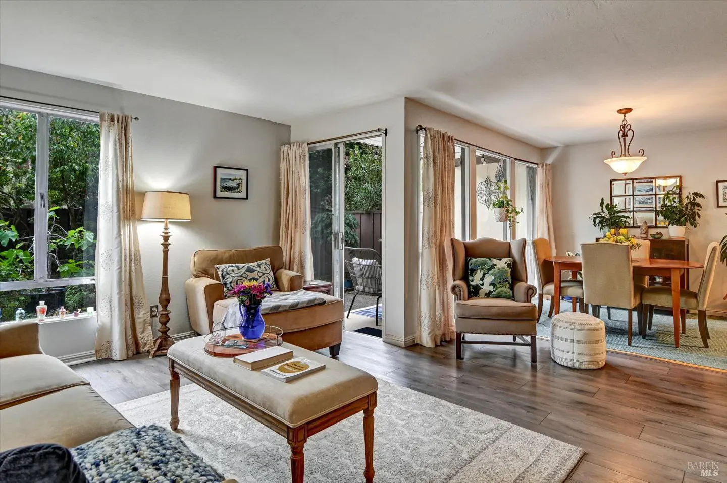 Living room with hardwood floors, beige walls, and natural light. Brown armchairs, a beige ottoman, and a dining table are visible.