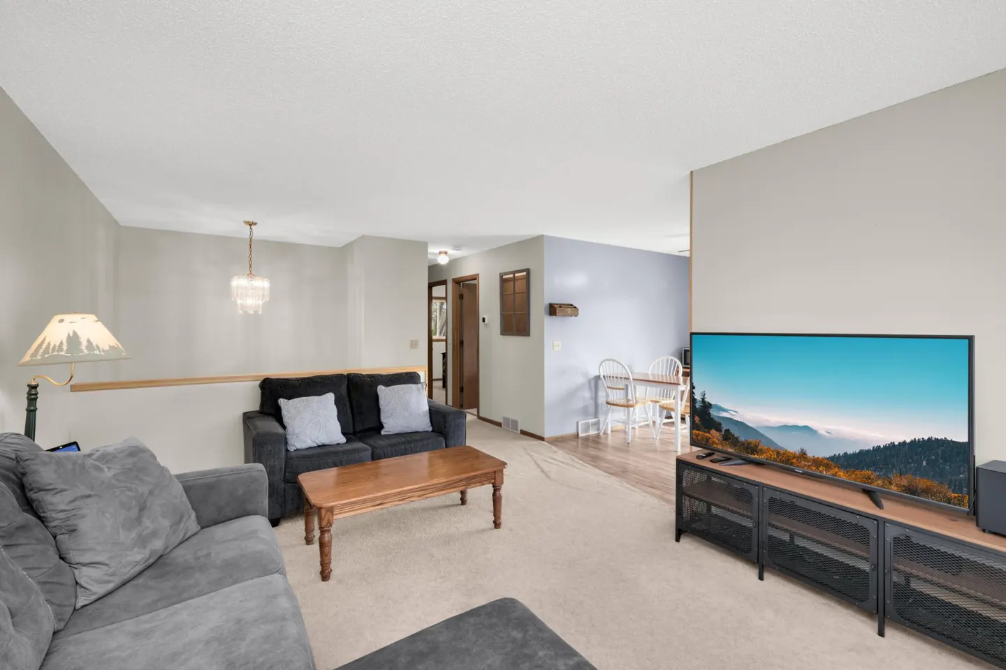 Living room with gray sofas, wood coffee table, and TV showing a mountain scene. Dining area with white table and chairs in the background.
