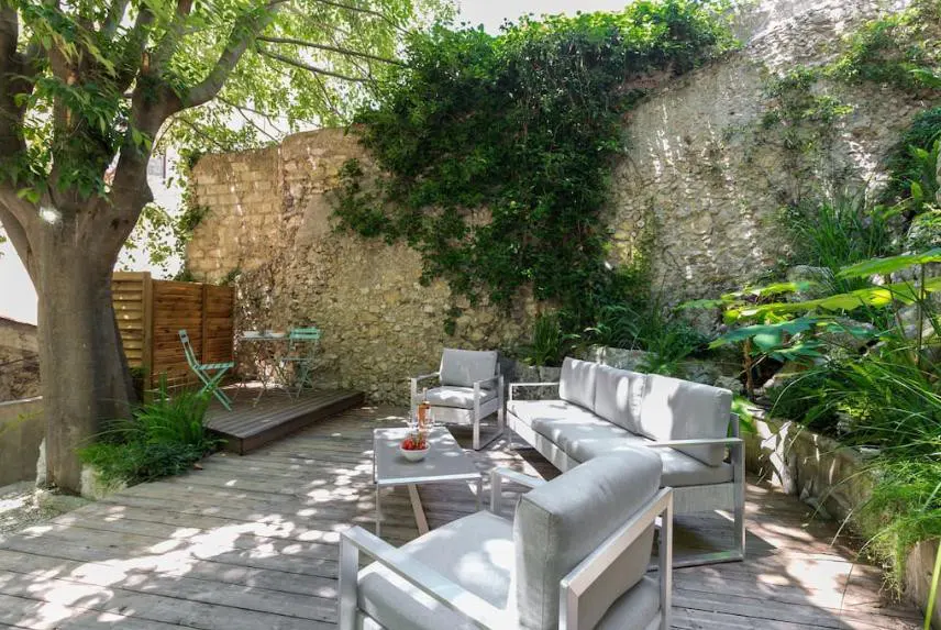 Outdoor patio with gray furniture on a wooden deck, stone wall backdrop with green foliage, and a large tree providing shade.