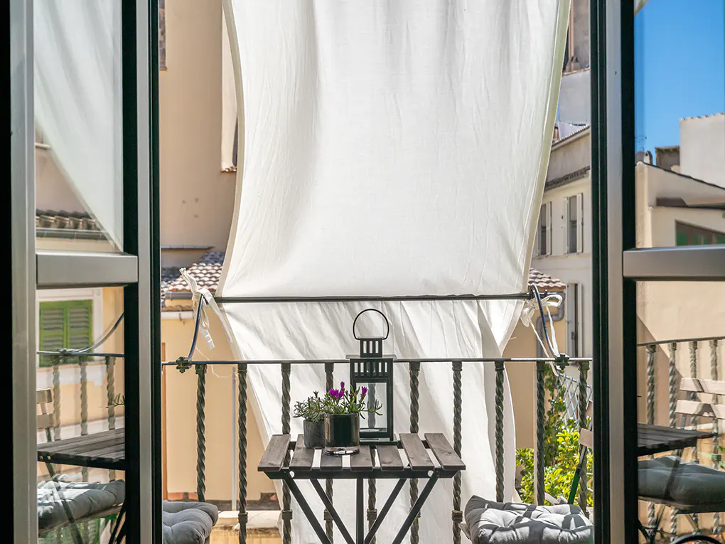 Balcony view through open doors. A small table with plants and a lantern sits under a white sunshade. Buildings are visible in the background.