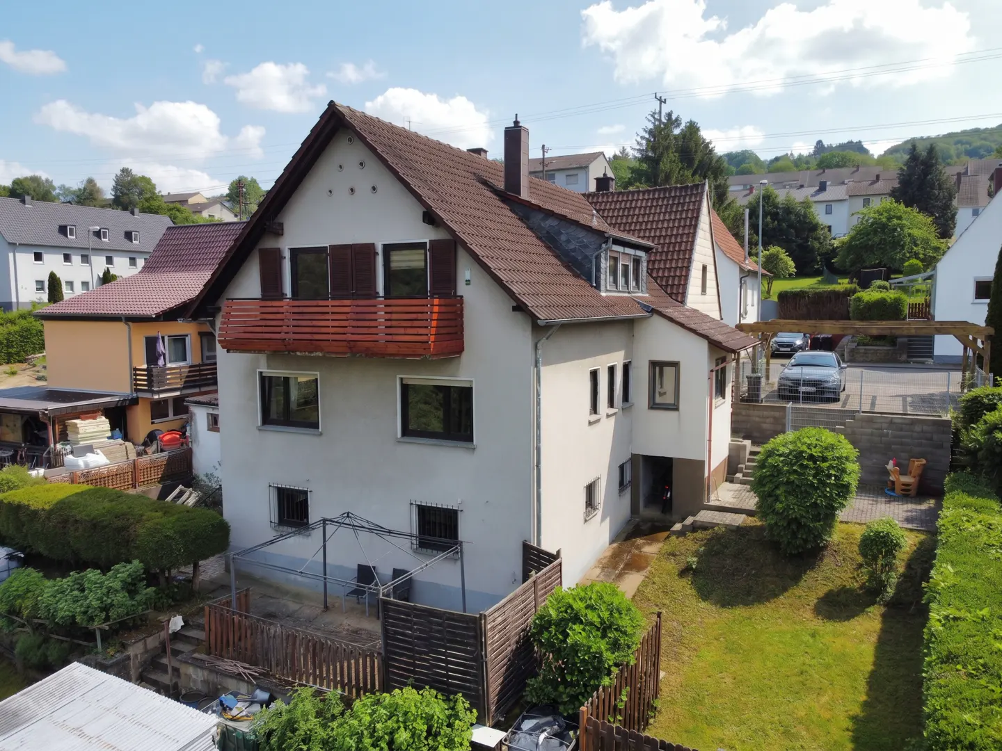 Exterior view of a multi-story beige house with a brown tiled roof, wooden balcony, and a backyard with a lawn and patio.
