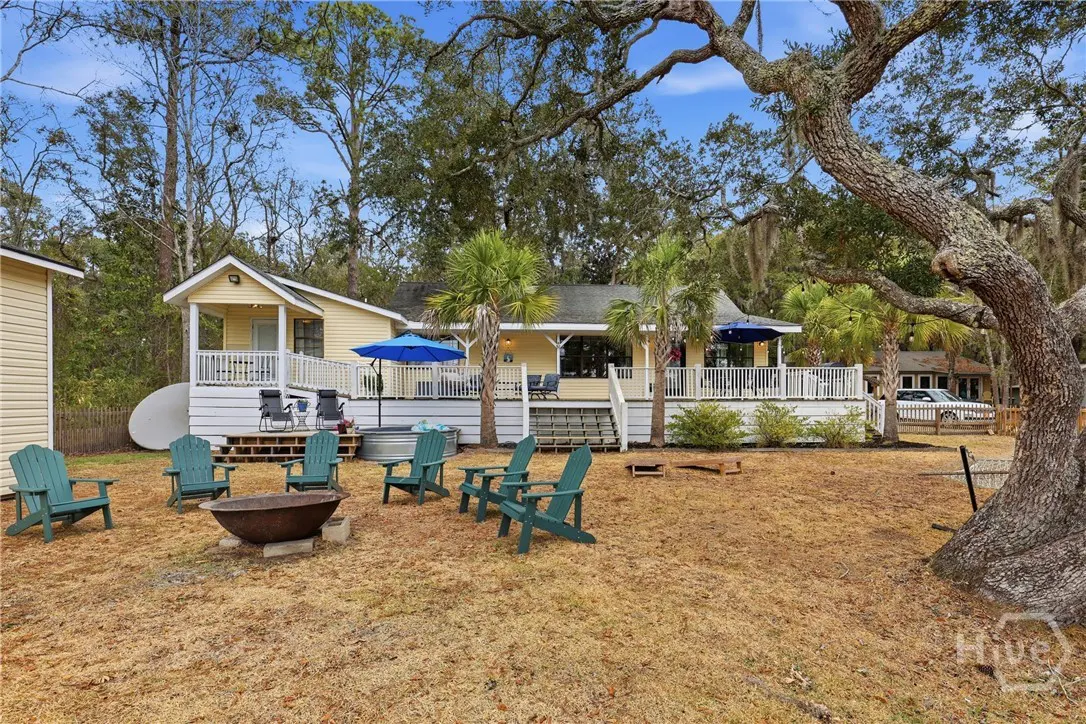 Backyard view of a yellow house with a white deck, blue umbrellas, and green Adirondack chairs around a fire pit.