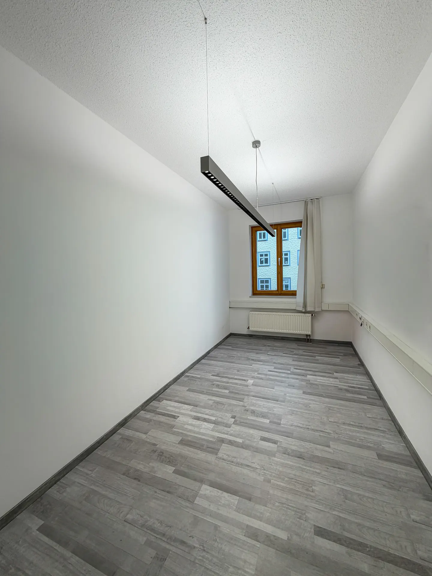 Empty room with gray wood-look floors, white walls, and a window. A modern, linear light fixture hangs from the ceiling.