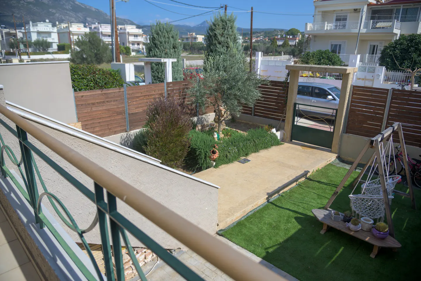 View of a fenced-in yard with a garden, concrete patio, artificial grass, and a swing set. A car is visible through the gate.