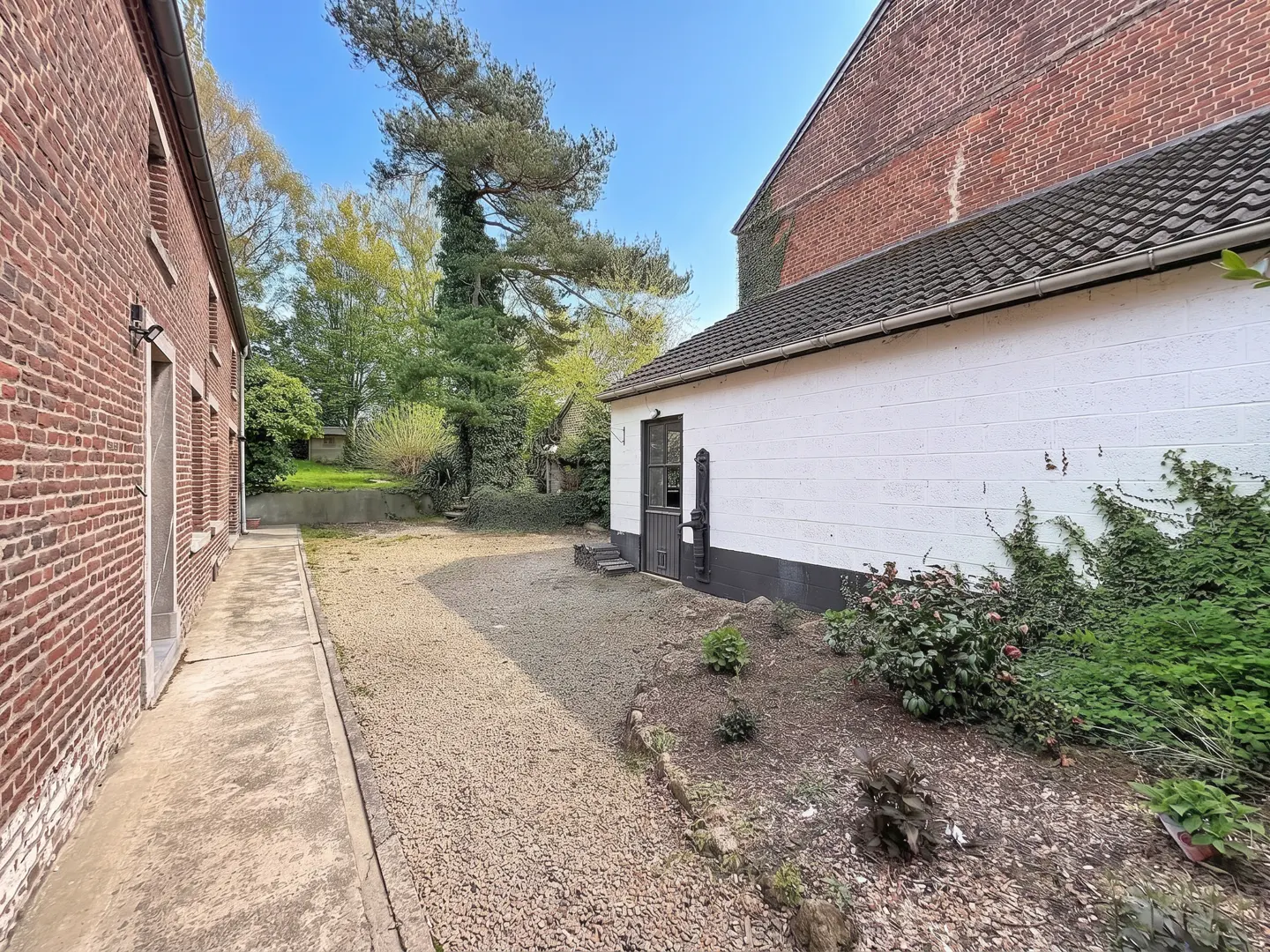 Exterior view of a red brick building with a white brick addition, gravel yard, and green trees.