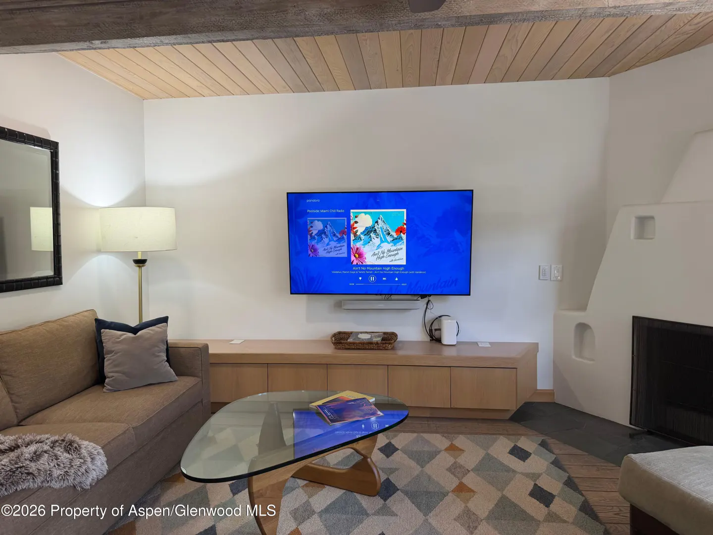 Living room with a brown sofa, glass coffee table, wall-mounted TV, and a white fireplace under a wood-paneled ceiling.