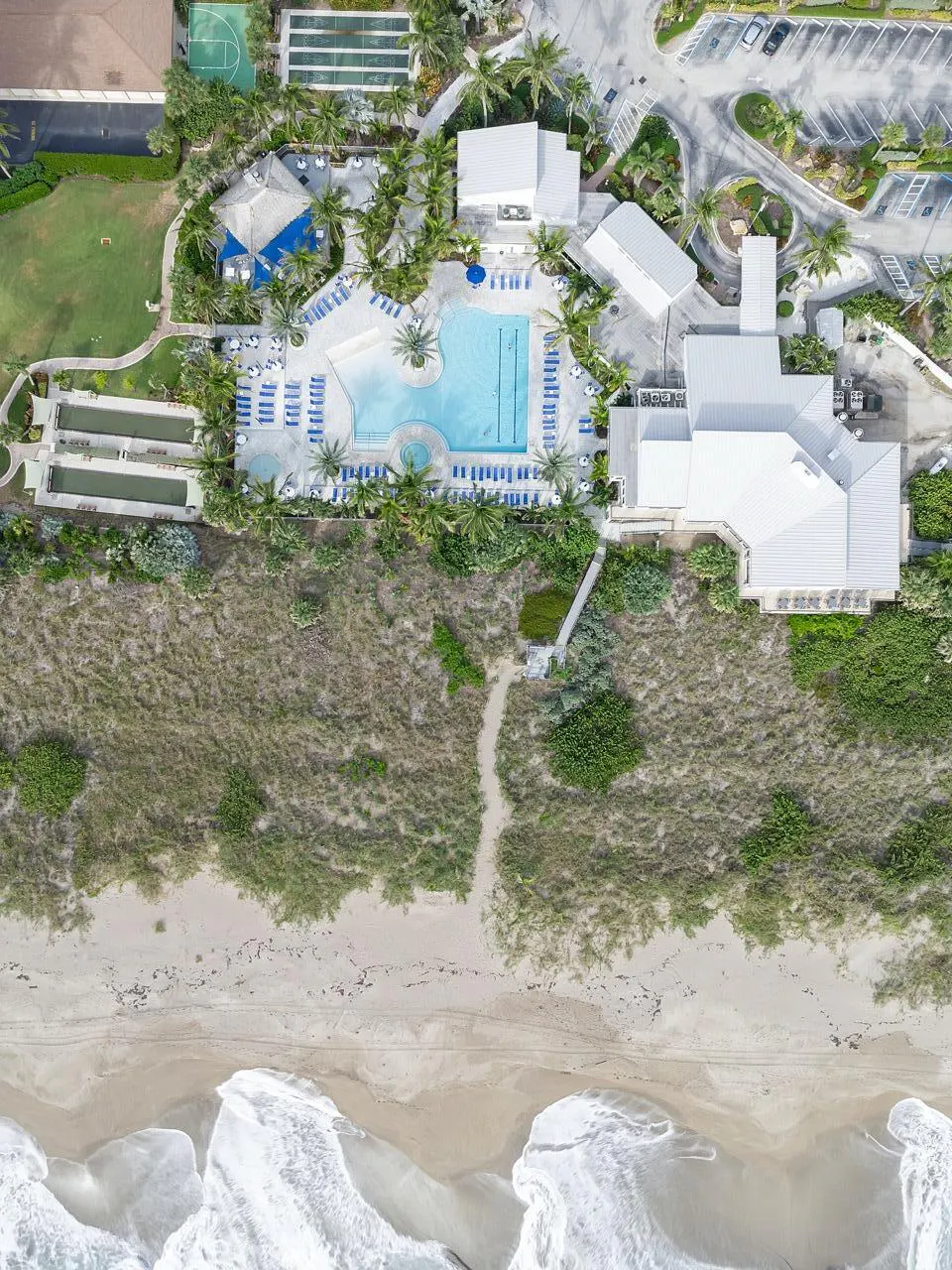 Aerial view of a beach resort with a pool, tennis courts, and a sandy beach with waves.
