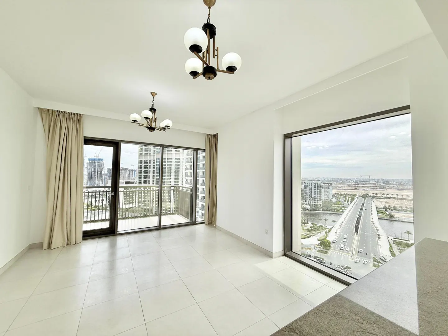 Bright, empty room with white walls, tile floor, and two chandeliers. Large windows offer city views with a bridge and buildings. Beige curtains frame a balcony door.
