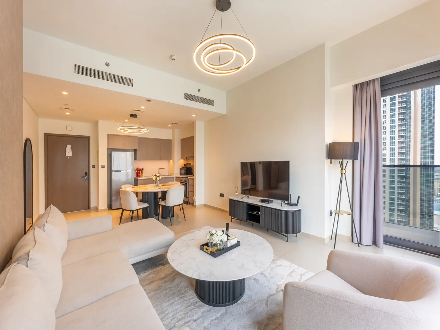 Modern open-plan living room with beige sofa, marble coffee table, TV, and dining area with kitchen in the background.
