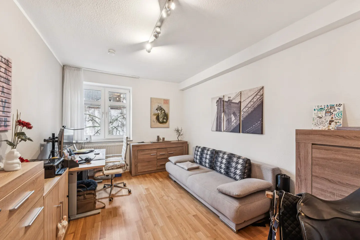 A bright living room with hardwood floors, a gray sofa with houndstooth pillows, and a desk by a window. Artwork adorns the white walls.