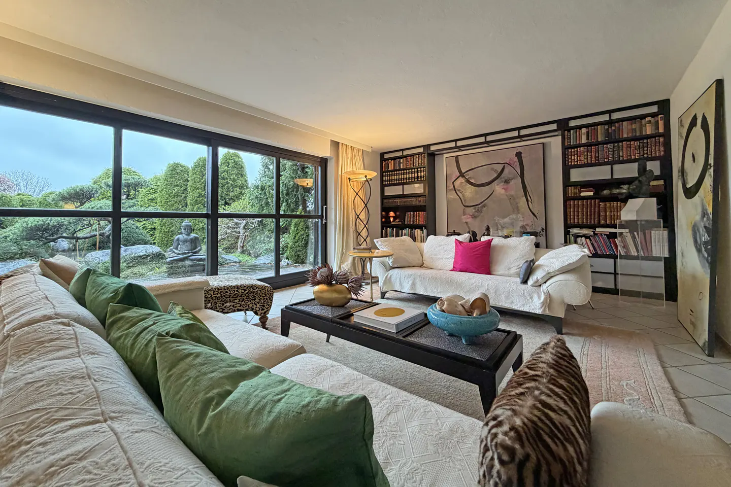 Living room with white sofas, green pillows, and a black coffee table. Large windows overlook a garden with a Buddha statue. Bookshelves line the back wall.