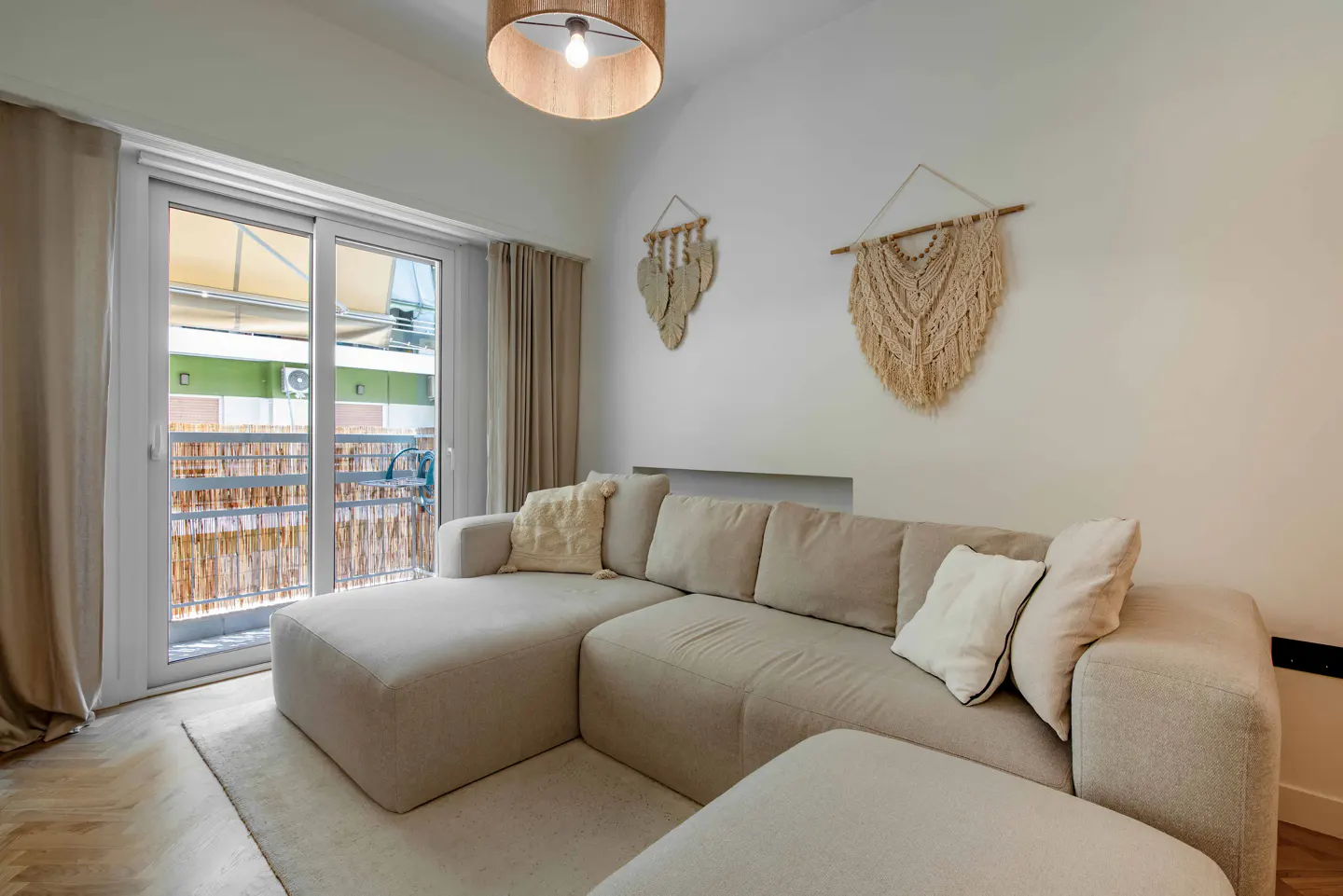 Living room with a beige sectional sofa, white pillows, and macrame wall hangings. Sliding glass doors lead to a balcony.