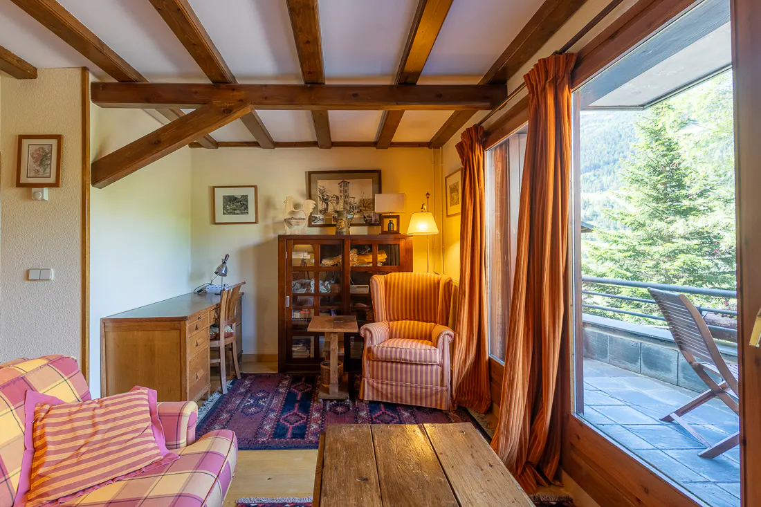 Cozy rustic room with exposed wooden beams, a striped armchair, a wooden desk, and a balcony view of trees.