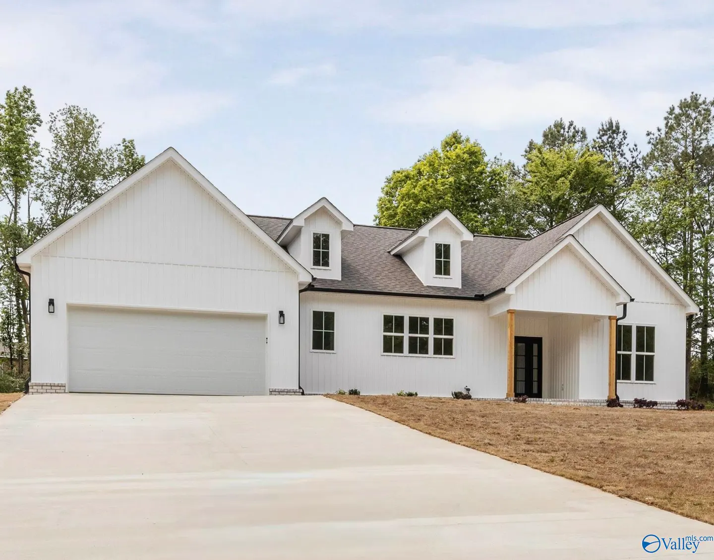 A modern farmhouse with white siding, a gray roof, and a concrete driveway. Trees are visible in the background.