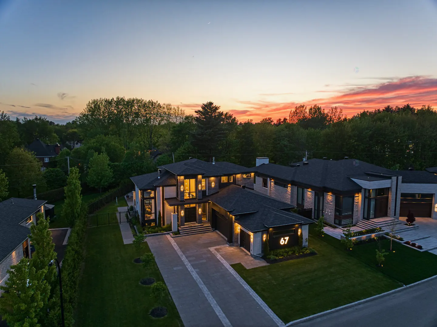Aerial view of a modern, gray stone house with black roof at dusk. Number 67 is visible on the garage. Trees and a colorful sky are in the background.