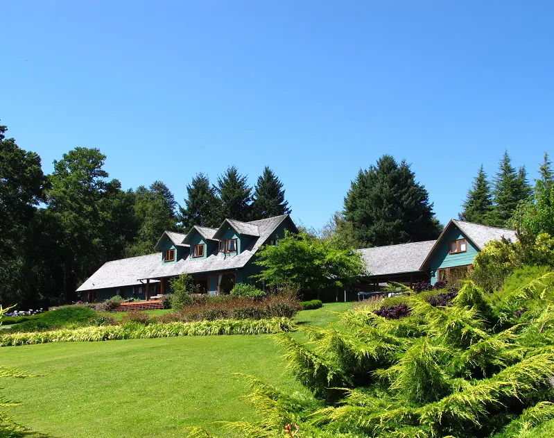 A long, teal house with a gray roof sits on a green lawn under a clear blue sky. Trees surround the house.