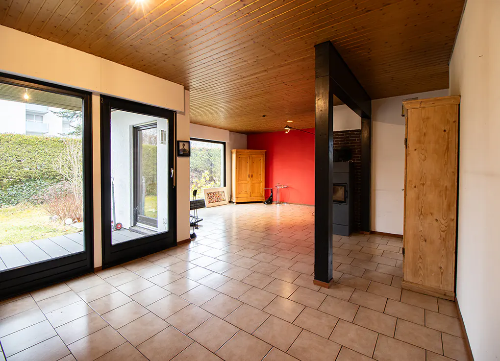 Bright living room with tile floor, wood ceiling, and black-framed sliding glass doors to a green yard. Red accent wall and wood cabinets.