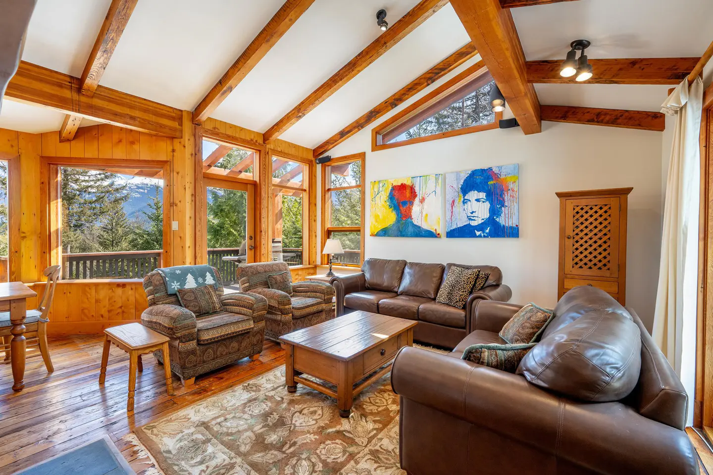 Living room with wood floors, exposed beams, and large windows with mountain views. Brown leather sofas and patterned armchairs surround a wooden coffee table.