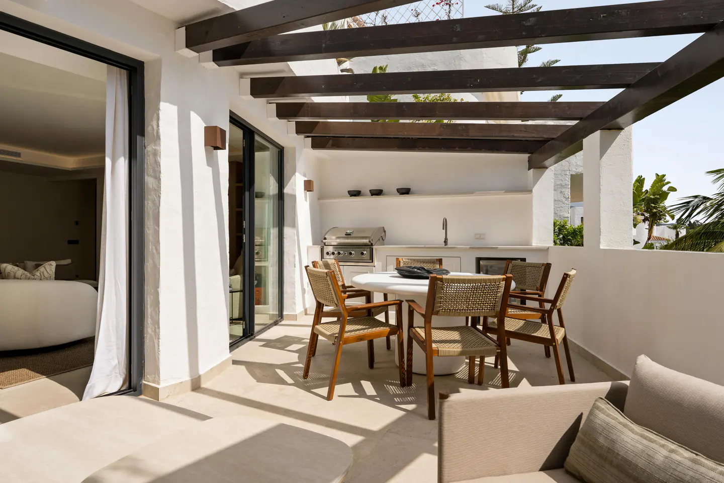 Outdoor patio with white walls, dark wood beams, and a built-in grill. A round table with six chairs sits in the center.