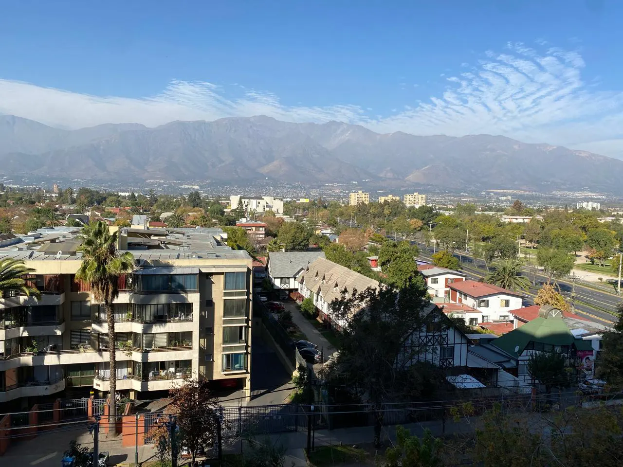 Cityscape view with buildings, trees, and mountains in the background under a blue sky with light clouds.