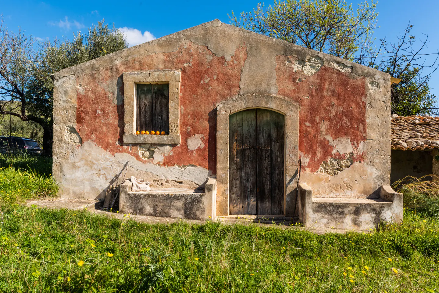 Rustic, weathered building with a red and beige facade, a dark wooden door, and oranges in the window, set in a grassy field.