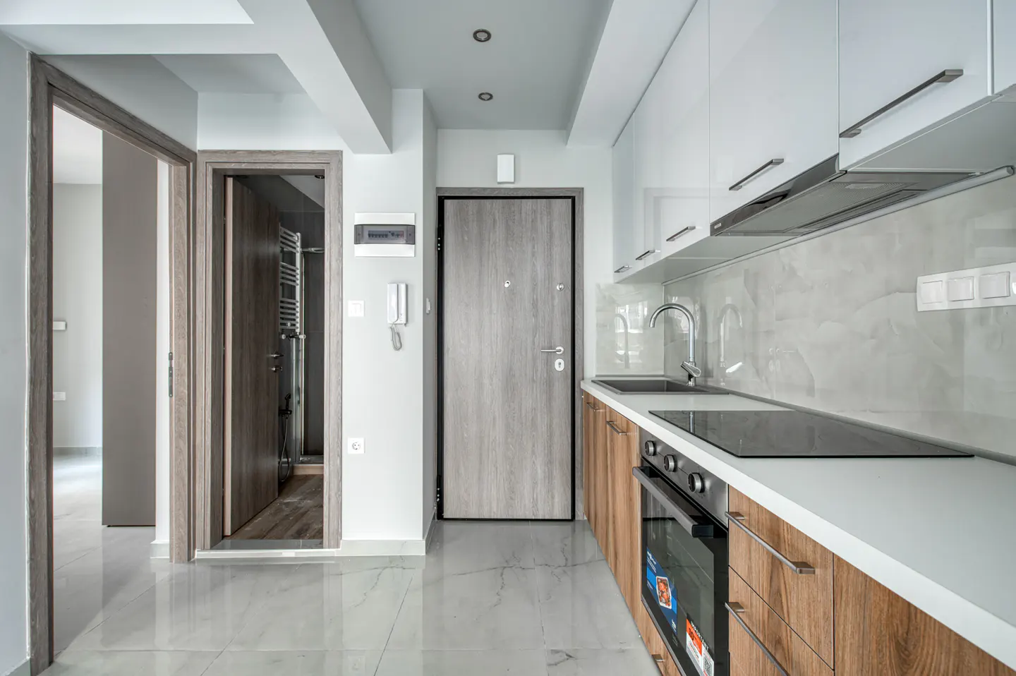 A modern kitchen with white cabinets, wood-grain lower cabinets, and marble floors. A gray door and open doorway are visible.