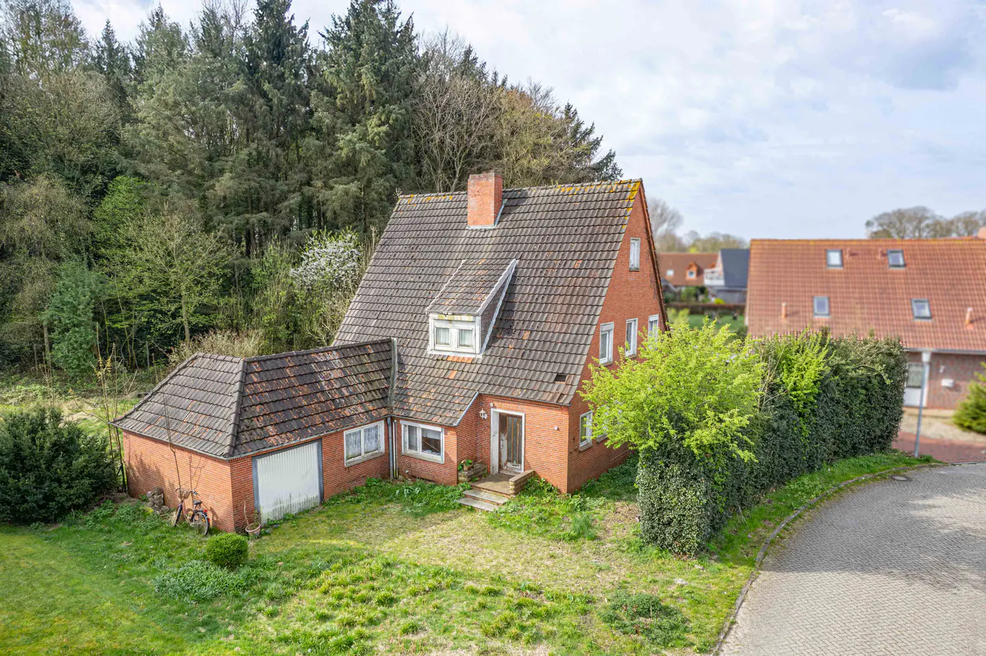 A red brick house with a tiled roof sits on a green lawn near a forest.
