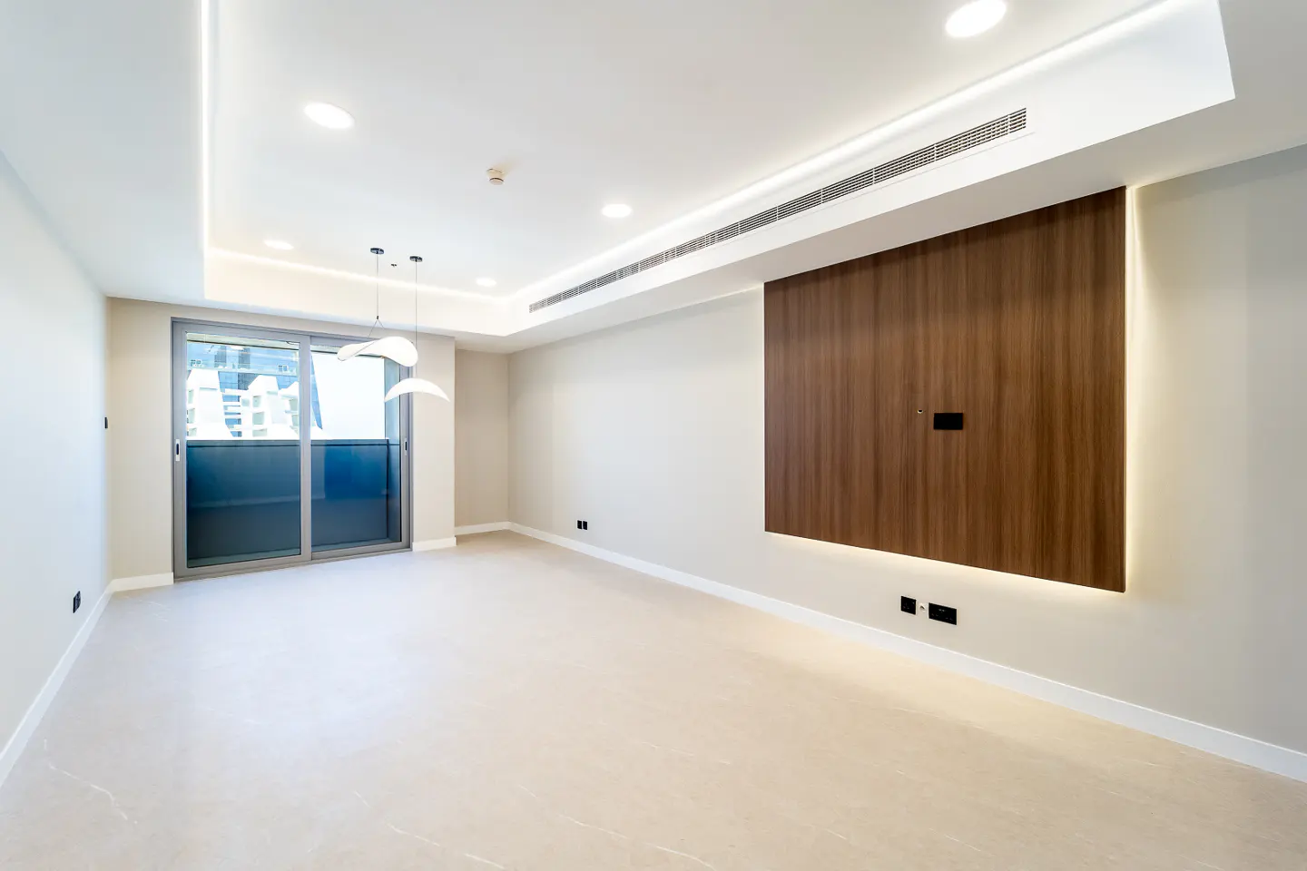 Bright, empty room with beige walls, tile floor, and a wood accent wall. A sliding glass door leads to a balcony. Modern pendant lights hang from the ceiling.