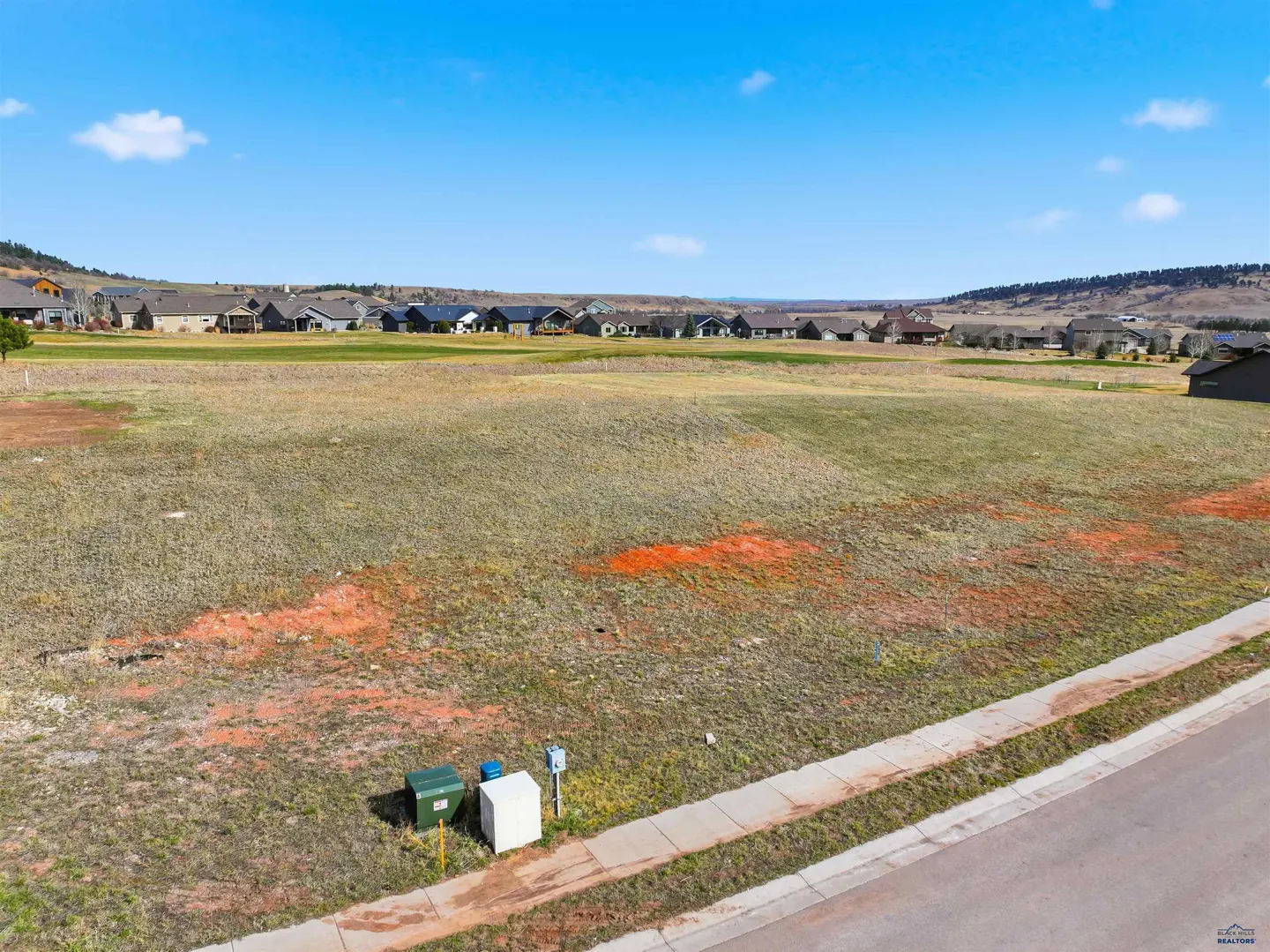 An aerial view of a vacant lot with utility boxes, a sidewalk, and houses in the background under a blue sky.