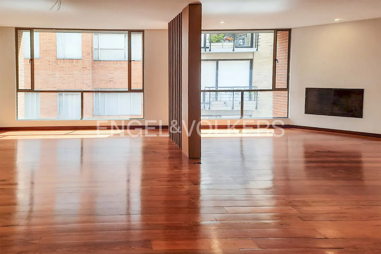 Empty room with shiny wood floors, large windows, and a wood slat divider. Brick buildings are visible through the windows.